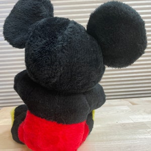 May include: A black plush Mickey Mouse toy sitting with its legs crossed. The toy has large black ears and is wearing red shorts with yellow trim.