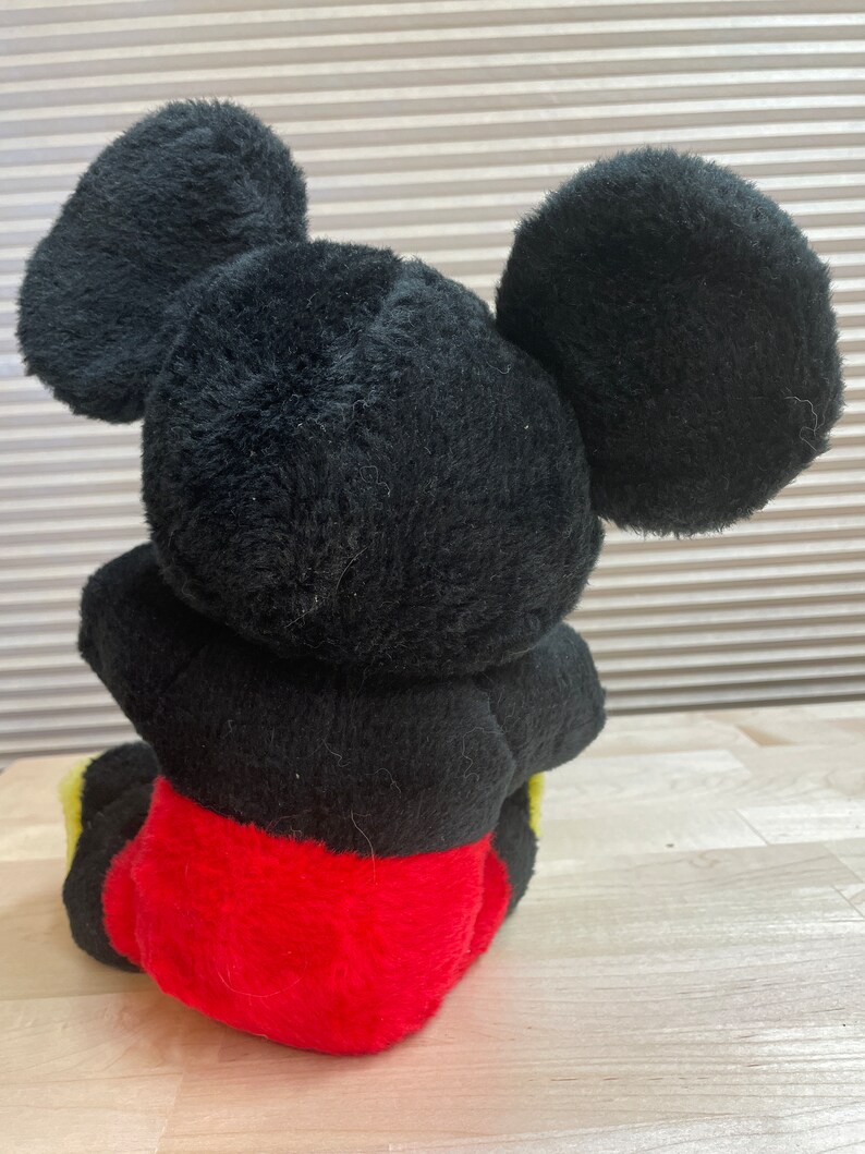 May include: A black plush Mickey Mouse toy sitting with its legs crossed. The toy has large black ears and is wearing red shorts with yellow trim.