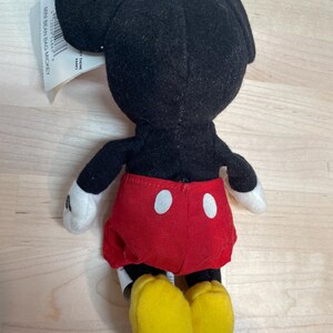 May include: A small Mickey Mouse plush toy, seen from the rear. The toy features a black head and body, red shorts with white buttons, white gloves, and yellow shoes. A tag is attached to the toy.