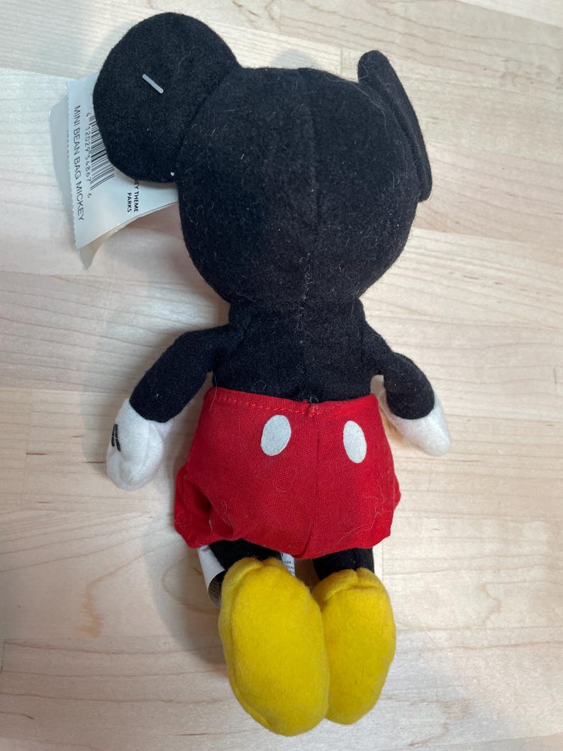 May include: A small Mickey Mouse plush toy, seen from the rear. The toy features a black head and body, red shorts with white buttons, white gloves, and yellow shoes. A tag is attached to the toy.