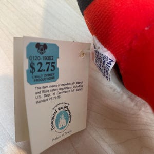 Puede incluir: Un juguete de peluche rojo, negro y amarillo con una etiqueta. La etiqueta tiene el texto "0120-19052" y "$2.75". La etiqueta tambi&eacute;n incluye el texto "Walt Disney Productions", "Disneyland Walt Disney World" y "Product of Taiwan".