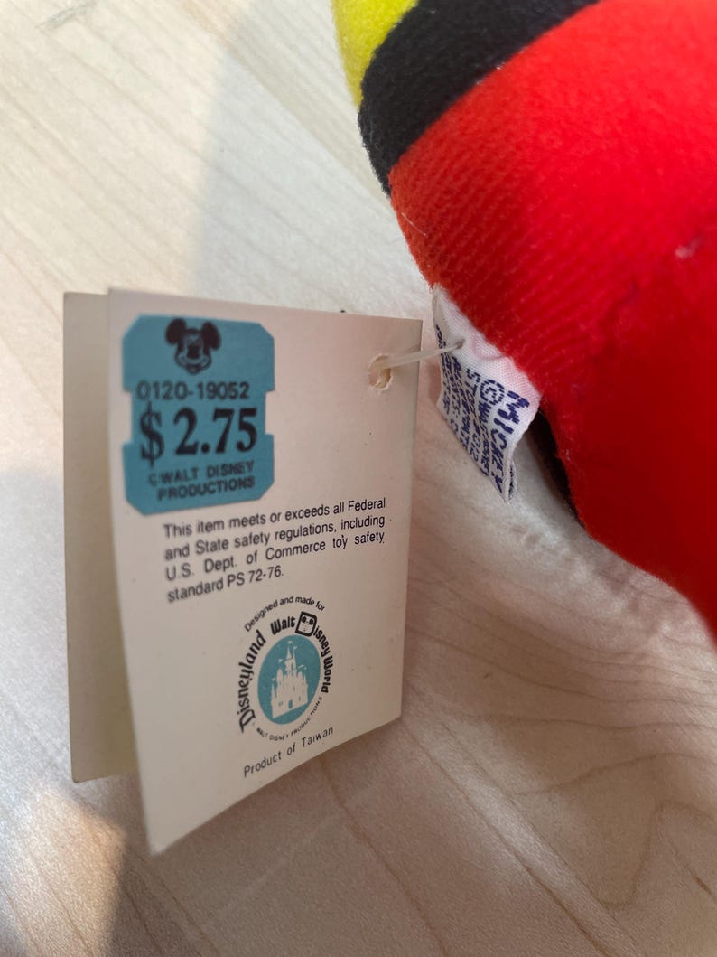 Puede incluir: Un juguete de peluche rojo, negro y amarillo con una etiqueta. La etiqueta tiene el texto "0120-19052" y "$2.75". La etiqueta tambi&eacute;n incluye el texto "Walt Disney Productions", "Disneyland Walt Disney World" y "Product of Taiwan".