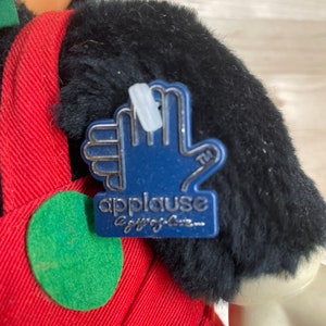 May include: A plush black and red Mickey Mouse toy with a blue "Applause" tag attached to its arm. The tag features a silver hand with the text "Applause a gift of love..."