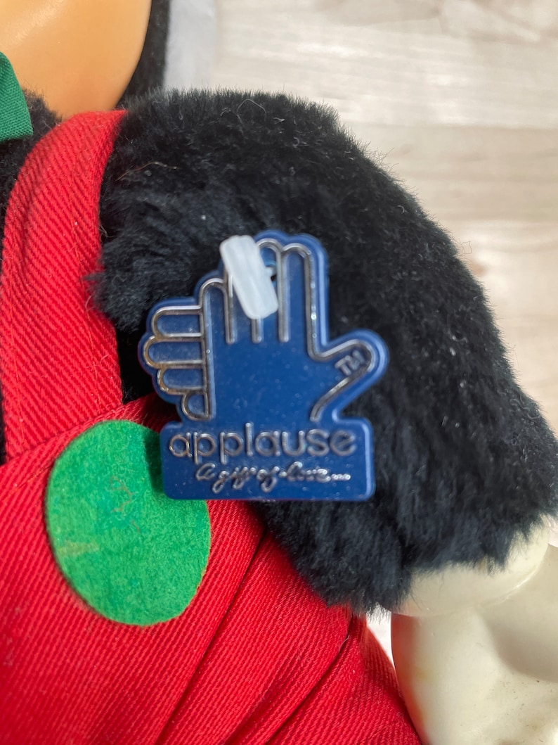 May include: A plush black and red Mickey Mouse toy with a blue "Applause" tag attached to its arm. The tag features a silver hand with the text "Applause a gift of love..."