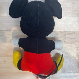 Puede incluir: Un peluche vintage de Mickey Mouse, visto desde atr&aacute;s. El peluche presenta orejas y cabeza negras, cuerpo negro, pantalones cortos rojos, zapatos amarillos y guantes blancos. Una etiqueta est&aacute; adjunta al juguete.