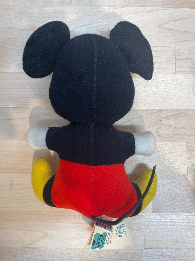 Puede incluir: Un peluche vintage de Mickey Mouse, visto desde atr&aacute;s. El peluche presenta orejas y cabeza negras, cuerpo negro, pantalones cortos rojos, zapatos amarillos y guantes blancos. Una etiqueta est&aacute; adjunta al juguete.