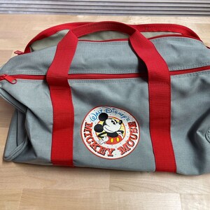 Puede incluir: Bolsa de lona gris con correas rojas y un logotipo de Mickey Mouse que dice "Walt Disney's Mickey Mouse".
