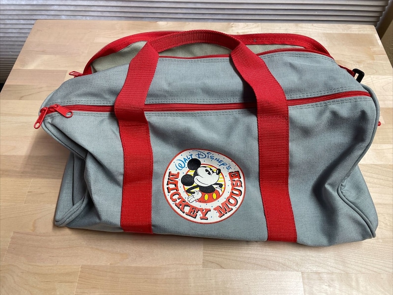 Puede incluir: Bolsa de lona gris con correas rojas y un logotipo de Mickey Mouse que dice "Walt Disney's Mickey Mouse".
