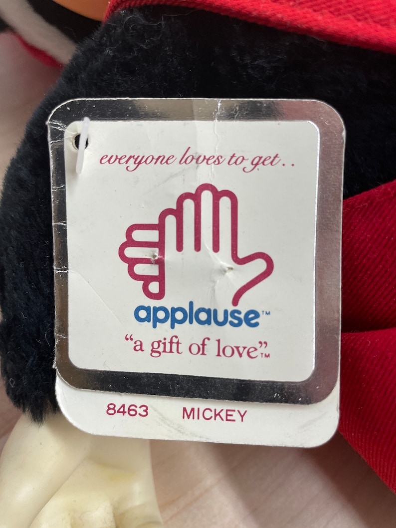 May include: A white tag with a silver border and the text "everyone loves to get... applause&trade; "a gift of love&trade;" 8463 MICKEY"  The tag features a red hand with the word "applause" in red text.