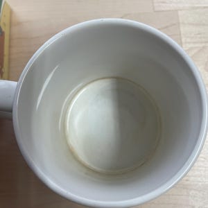 K&ouml;nnte beinhalten: Eine wei&szlig;e Keramik-Tasse mit Henkel, von oben betrachtet. Das Innere weist Gebrauchsspuren mit leichten Verf&auml;rbungen am Boden auf. Die Tasse steht auf einer hellen Holzoberfl&auml;che.