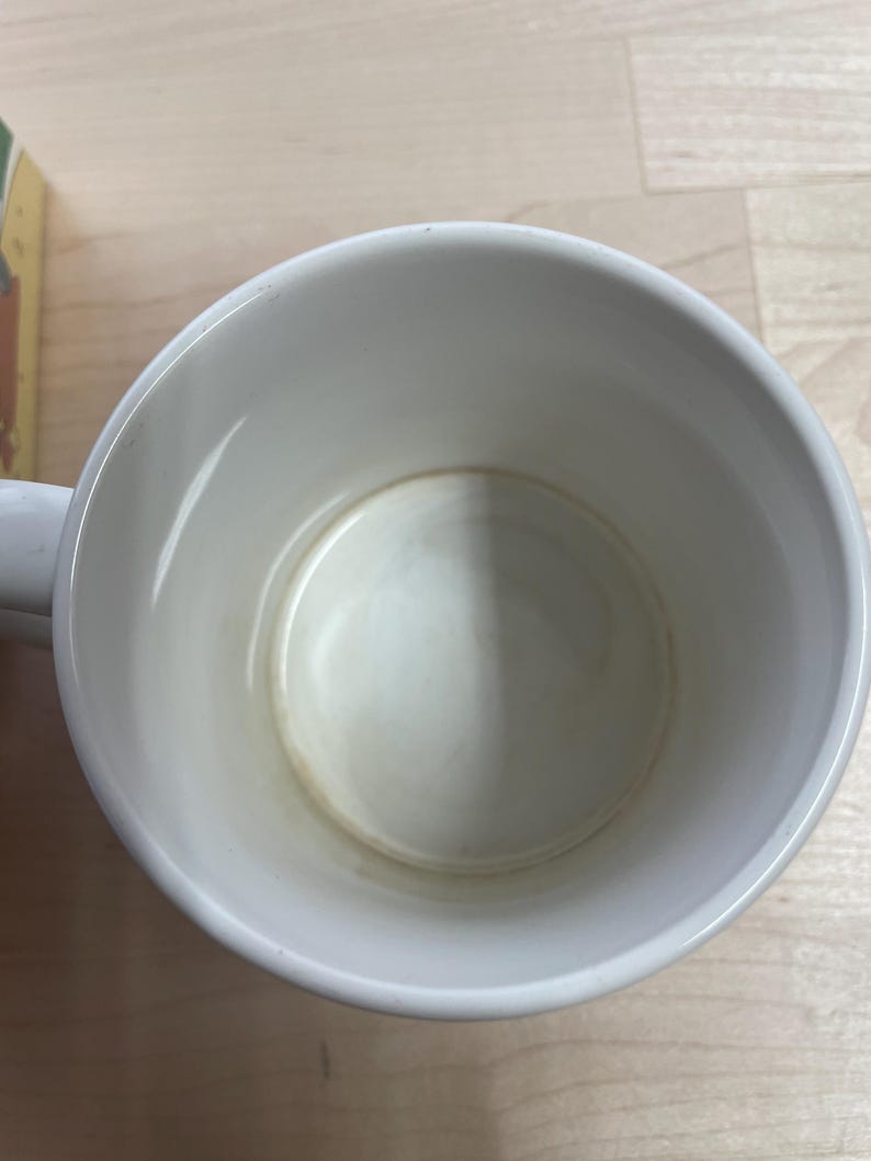 K&ouml;nnte beinhalten: Eine wei&szlig;e Keramik-Tasse mit Henkel, von oben betrachtet. Das Innere weist Gebrauchsspuren mit leichten Verf&auml;rbungen am Boden auf. Die Tasse steht auf einer hellen Holzoberfl&auml;che.
