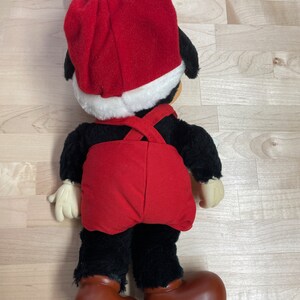 May include: A plush toy of Mickey Mouse wearing a red Santa hat and red overalls. The toy is standing on a wooden surface.