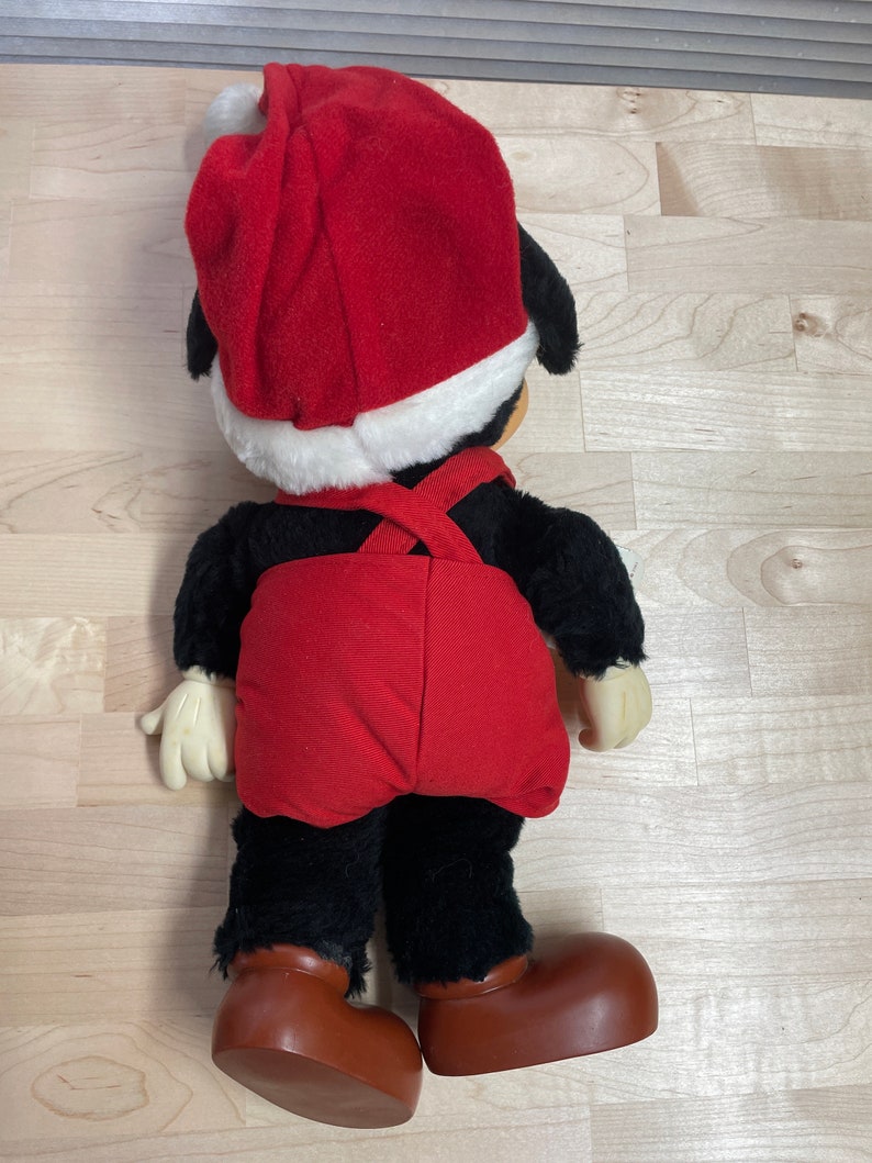May include: A plush toy of Mickey Mouse wearing a red Santa hat and red overalls. The toy is standing on a wooden surface.