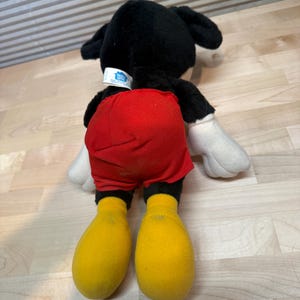 May include: A plush Mickey Mouse toy, viewed from the back, with black fur, red shorts, yellow feet, and white gloves. The toy has a tag on the back of its neck. The toy is lying on a light-coloured wooden surface.