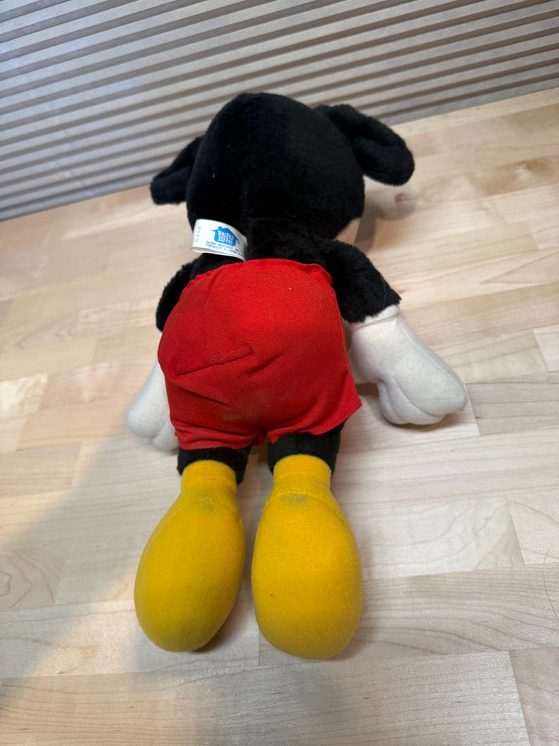 May include: A plush Mickey Mouse toy, viewed from the back, with black fur, red shorts, yellow feet, and white gloves. The toy has a tag on the back of its neck. The toy is lying on a light-coloured wooden surface.