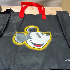 May include: A black reusable tote bag featuring a graphic of Mickey Mouse's face surrounded by a yellow spray-paint effect halo. The bag has two red fabric handles and the word Disney printed in white text in the bottom right corner.