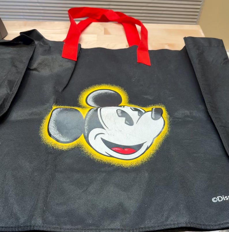 May include: A black reusable tote bag featuring a graphic of Mickey Mouse's face surrounded by a yellow spray-paint effect halo. The bag has two red fabric handles and the word Disney printed in white text in the bottom right corner.