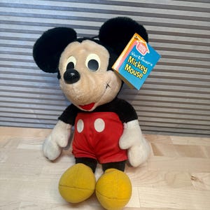 May include: A plush Mickey Mouse doll with black ears, a black body, red shorts with white buttons, and yellow feet. The doll has a tag that reads "Walt Disney's Mickey Mouse". The doll is sitting on a light-coloured surface.