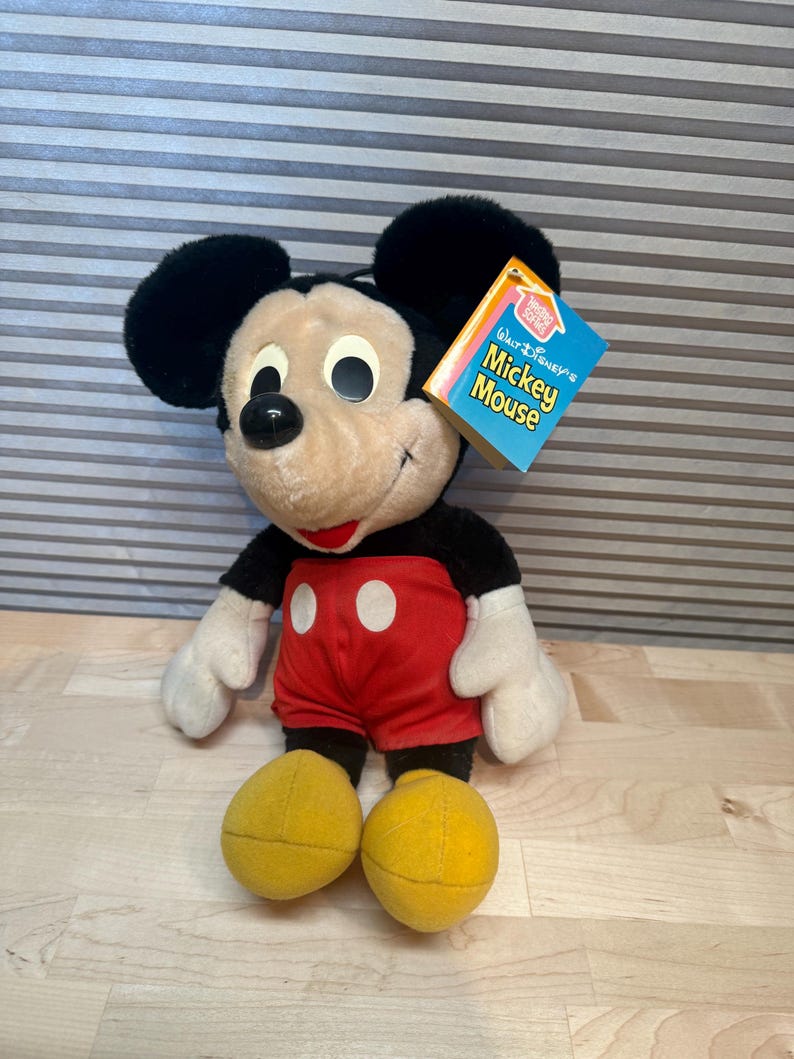 May include: A plush Mickey Mouse doll with black ears, a black body, red shorts with white buttons, and yellow feet. The doll has a tag that reads "Walt Disney's Mickey Mouse". The doll is sitting on a light-coloured surface.