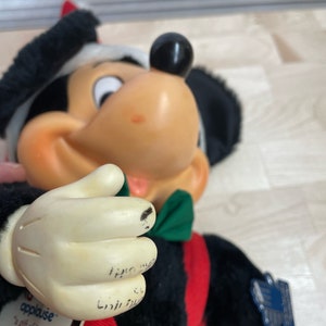 May include: A black and white plush Mickey Mouse wearing a red and green holiday outfit. The plush toy is holding a green bow tie in its hand. The tag on the toy reads "applause...a gift of love." and "Mickey".