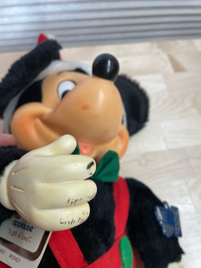 May include: A black and white plush Mickey Mouse wearing a red and green holiday outfit. The plush toy is holding a green bow tie in its hand. The tag on the toy reads "applause...a gift of love." and "Mickey".
