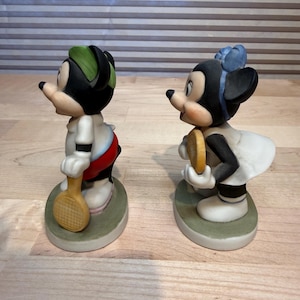Puede incluir: Dos figuras de cer&aacute;mica de ratones de dibujos animados, uno masculino y otro femenino, representados con atuendo de tenis. El rat&oacute;n masculino lleva una gorra verde y sostiene una raqueta de tenis. La ratona lleva un lazo azul y sostiene una pelota de tenis.