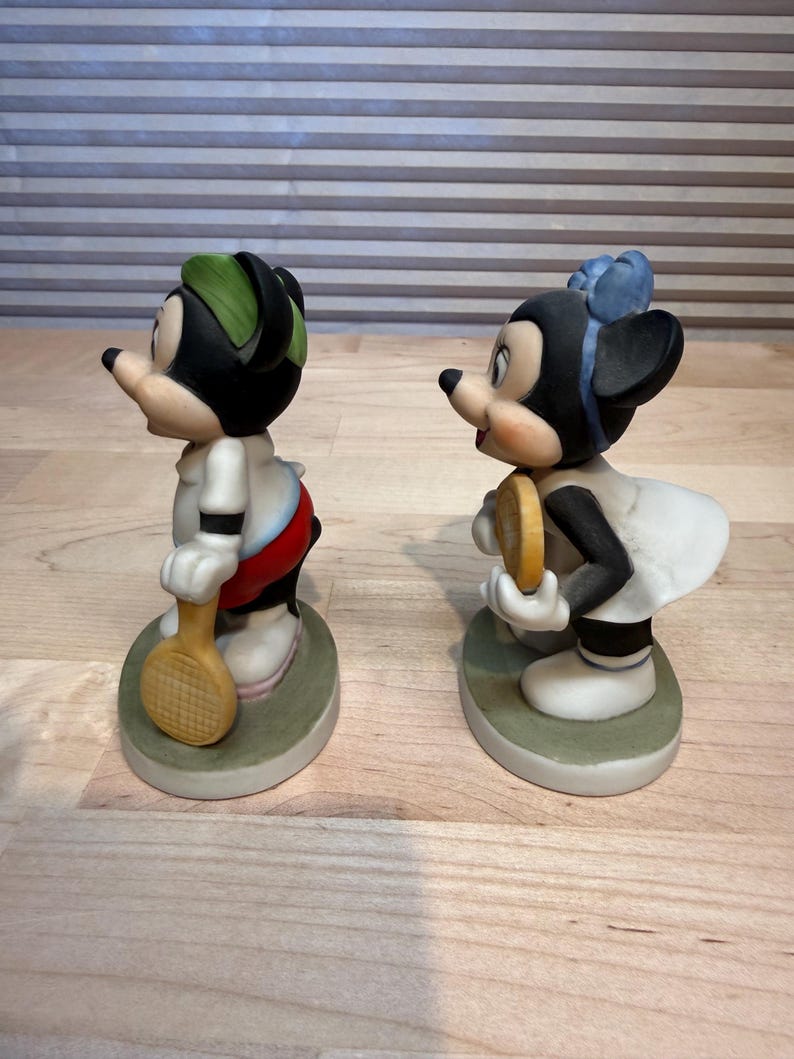 Puede incluir: Dos figuras de cer&aacute;mica de ratones de dibujos animados, uno masculino y otro femenino, representados con atuendo de tenis. El rat&oacute;n masculino lleva una gorra verde y sostiene una raqueta de tenis. La ratona lleva un lazo azul y sostiene una pelota de tenis.