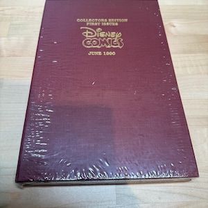 May include: A sealed, burgundy-coloured "Disney Comics" collector's edition from June 1990. The cover features gold lettering and is wrapped in clear plastic. The comic book is lying on a light-coloured wooden surface.