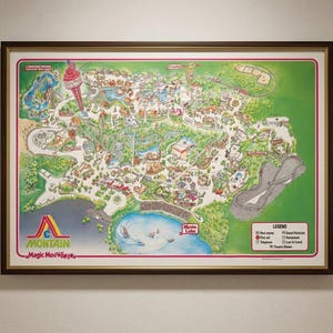May include: A framed vintage map of Magic Mountain amusement park featuring a green landscape, a blue lake labeled Monte Lake, a tall red tower, and various ride illustrations. The bottom left corner displays the Magic Mountain logo and text.