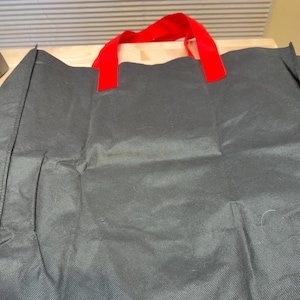 May include: A black reusable tote bag featuring a textured, non-woven fabric construction. The bag includes two bright red fabric handles securely attached to the top edge, providing a clear contrast against the dark, matte black body of the bag.