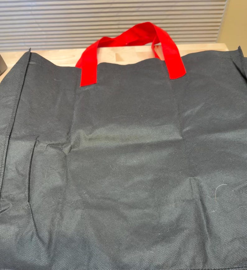May include: A black reusable tote bag featuring a textured, non-woven fabric construction. The bag includes two bright red fabric handles securely attached to the top edge, providing a clear contrast against the dark, matte black body of the bag.