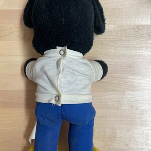 May include: Black plush toy dog wearing a white shirt and blue trousers. The dog has yellow feet and black ears.