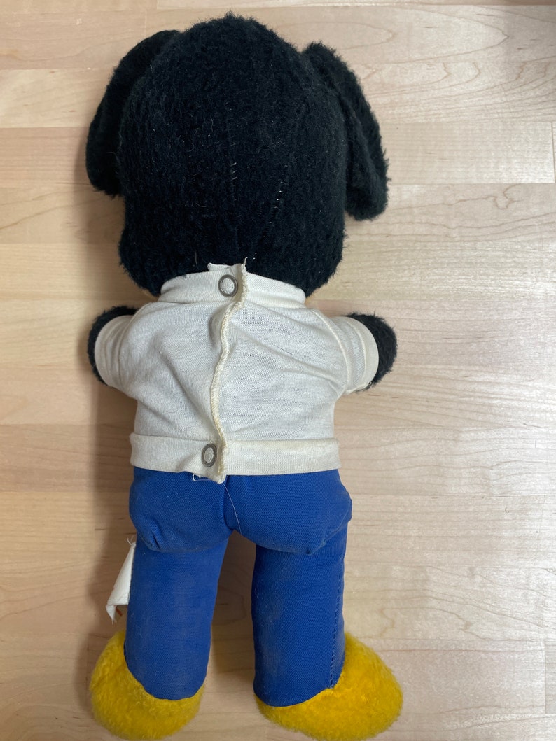 May include: Black plush toy dog wearing a white shirt and blue trousers. The dog has yellow feet and black ears.