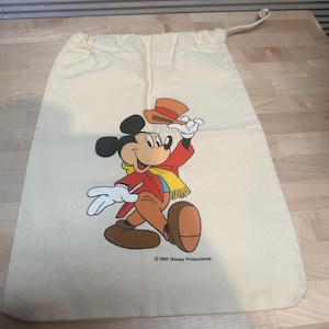 May include: Cream-colored drawstring bag featuring a colorful illustration of Mickey Mouse in a red jacket, brown pants, and a yellow scarf, holding a hat. The bag has a rope drawstring closure and the text "Walt Disney Productions".
