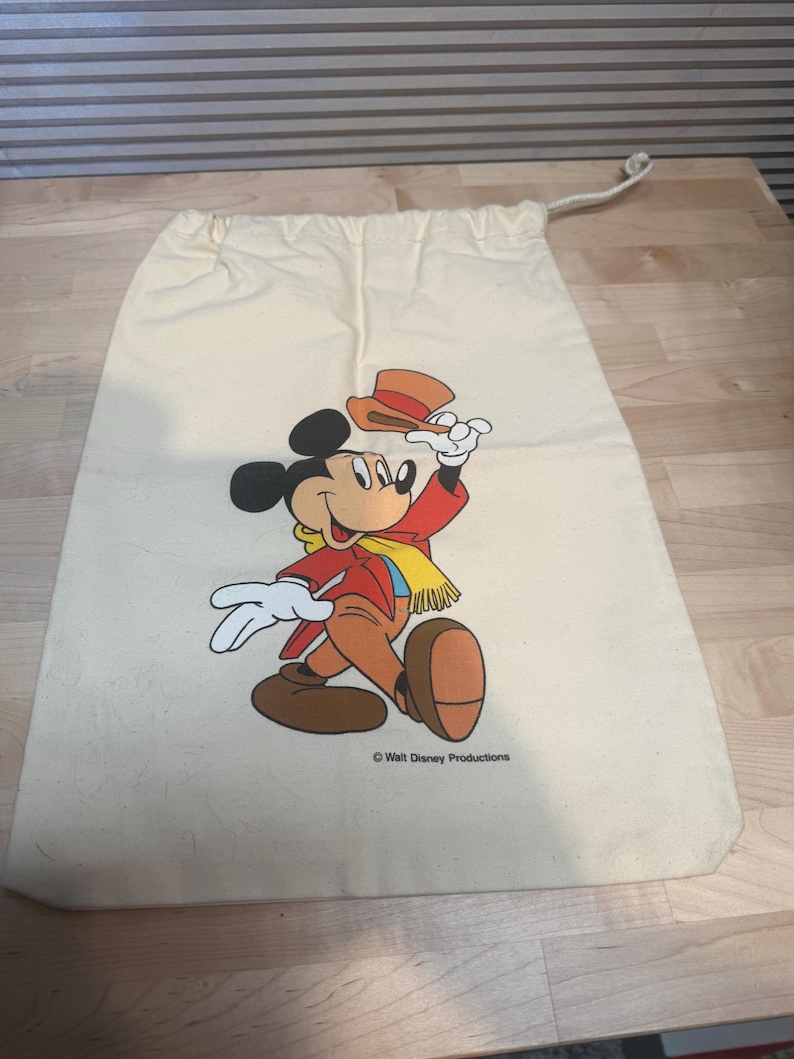 May include: Cream-colored drawstring bag featuring a colorful illustration of Mickey Mouse in a red jacket, brown pants, and a yellow scarf, holding a hat. The bag has a rope drawstring closure and the text "Walt Disney Productions".