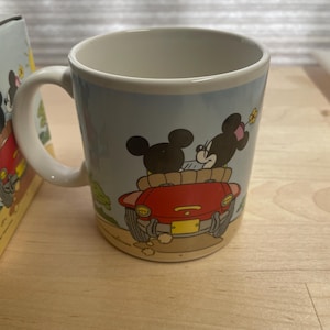 K&ouml;nnte beinhalten: Eine Keramik-Kaffeetasse mit einer Cartoon-Illustration von Mickey und Minnie Mouse in einem roten Auto. Die Tasse hat einen wei&szlig;en Henkel und einen hellblauen Hintergrund mit einer Landschaftsszene. Die Tasse ist wahrscheinlich ein Sammlerst&uuml;ck.