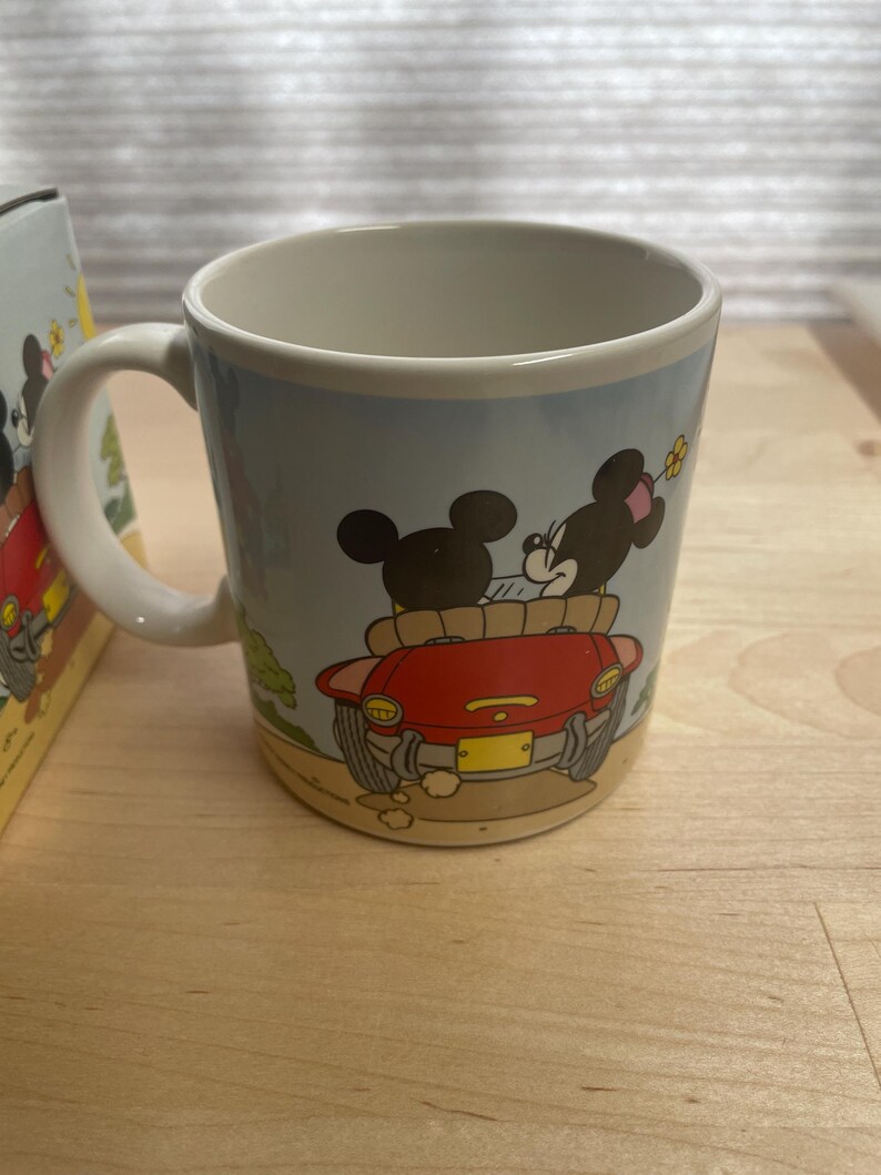 K&ouml;nnte beinhalten: Eine Keramik-Kaffeetasse mit einer Cartoon-Illustration von Mickey und Minnie Mouse in einem roten Auto. Die Tasse hat einen wei&szlig;en Henkel und einen hellblauen Hintergrund mit einer Landschaftsszene. Die Tasse ist wahrscheinlich ein Sammlerst&uuml;ck.