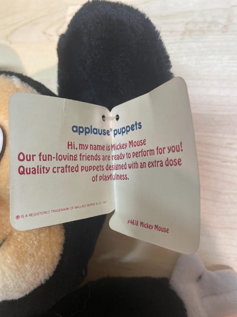 May include: A Mickey Mouse puppet with a black head and tan face. The puppet has a tag that reads "applause puppets" and "Hi, my name is Mickey Mouse." The tag also includes the text "Quality crafted puppets designed with an extra dose of playfulness."