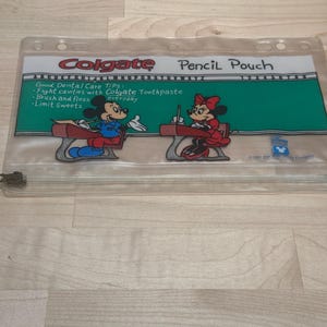 Peut inclure: Trousse à crayons transparente avec des illustrations de Mickey et Minnie Mouse. La trousse présente un design de tableau noir vert avec le logo Colgate et des conseils de soins dentaires. Le texte sur la trousse indique "Pencil Pouch" et "Colgate".