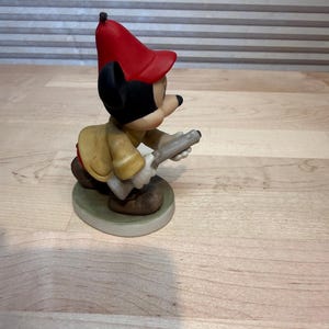 May include: A ceramic figurine of a cartoon character wearing a red hat and yellow jacket, holding a gray object. The figurine is on a round, green-edged base. The character has black ears and a black nose.