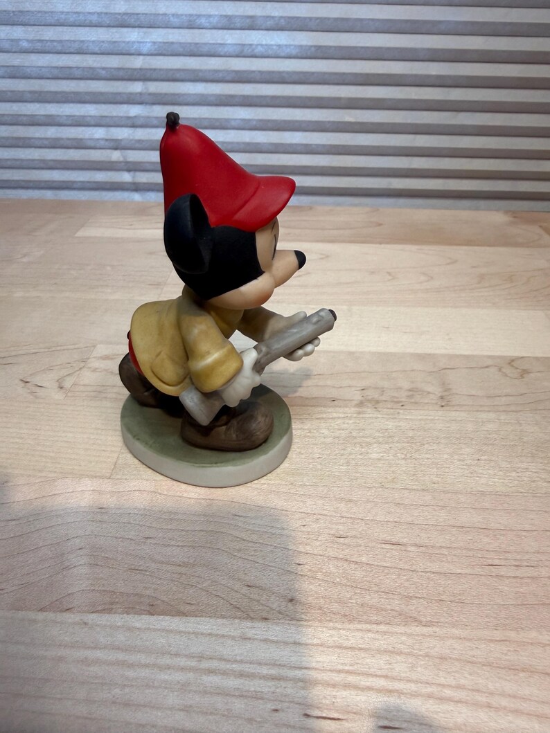 May include: A ceramic figurine of a cartoon character wearing a red hat and yellow jacket, holding a gray object. The figurine is on a round, green-edged base. The character has black ears and a black nose.