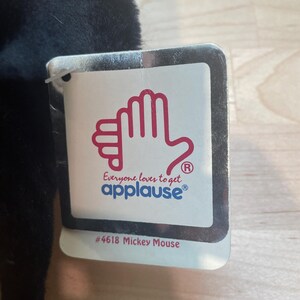 May include: A black plush toy with a tag that reads "Everyone loves to get applause&reg;" and "#4618 Mickey Mouse". The tag features a red hand design. The toy is partially visible, with a soft, textured appearance.