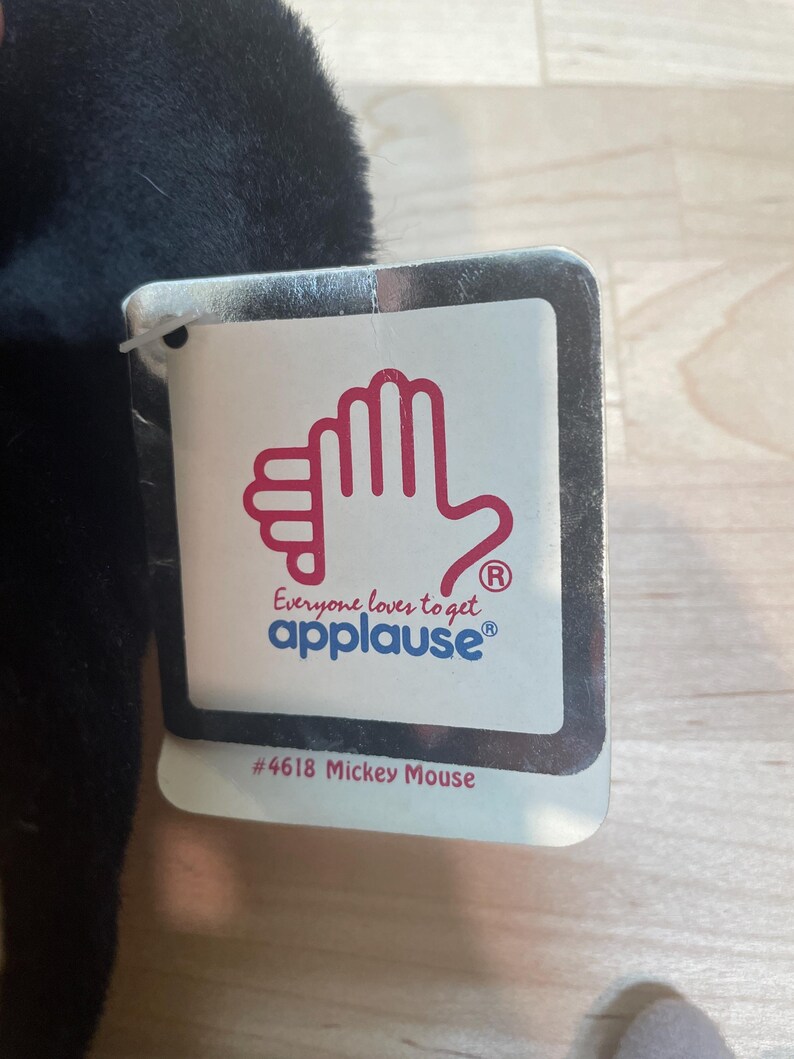 May include: A black plush toy with a tag that reads "Everyone loves to get applause&reg;" and "#4618 Mickey Mouse". The tag features a red hand design. The toy is partially visible, with a soft, textured appearance.