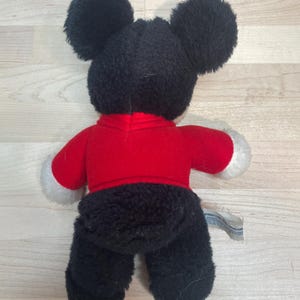 Puede incluir: Un peluche de un rat&oacute;n de dibujos animados, visto desde atr&aacute;s. El rat&oacute;n tiene grandes orejas negras, una camisa roja con pu&ntilde;os blancos y pantalones negros. Una peque&ntilde;a etiqueta est&aacute; adherida al juguete.