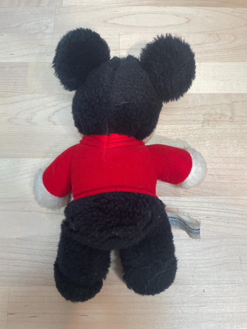 Puede incluir: Un peluche de un rat&oacute;n de dibujos animados, visto desde atr&aacute;s. El rat&oacute;n tiene grandes orejas negras, una camisa roja con pu&ntilde;os blancos y pantalones negros. Una peque&ntilde;a etiqueta est&aacute; adherida al juguete.
