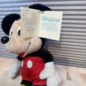 May include: A vintage Mickey Mouse soft toy, with a black head and ears, a cream-coloured face, red shorts with white buttons, and yellow feet. The toy has a tag with text and signatures.