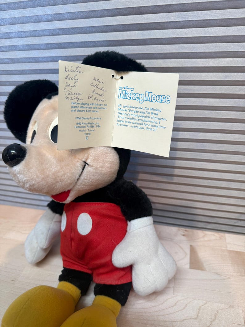 May include: A vintage Mickey Mouse soft toy, with a black head and ears, a cream-coloured face, red shorts with white buttons, and yellow feet. The toy has a tag with text and signatures.