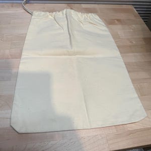 May include: A cream-colored drawstring bag made of canvas fabric. The bag is rectangular with a rounded bottom and a drawstring closure at the top. The bag is laying on a wooden surface.