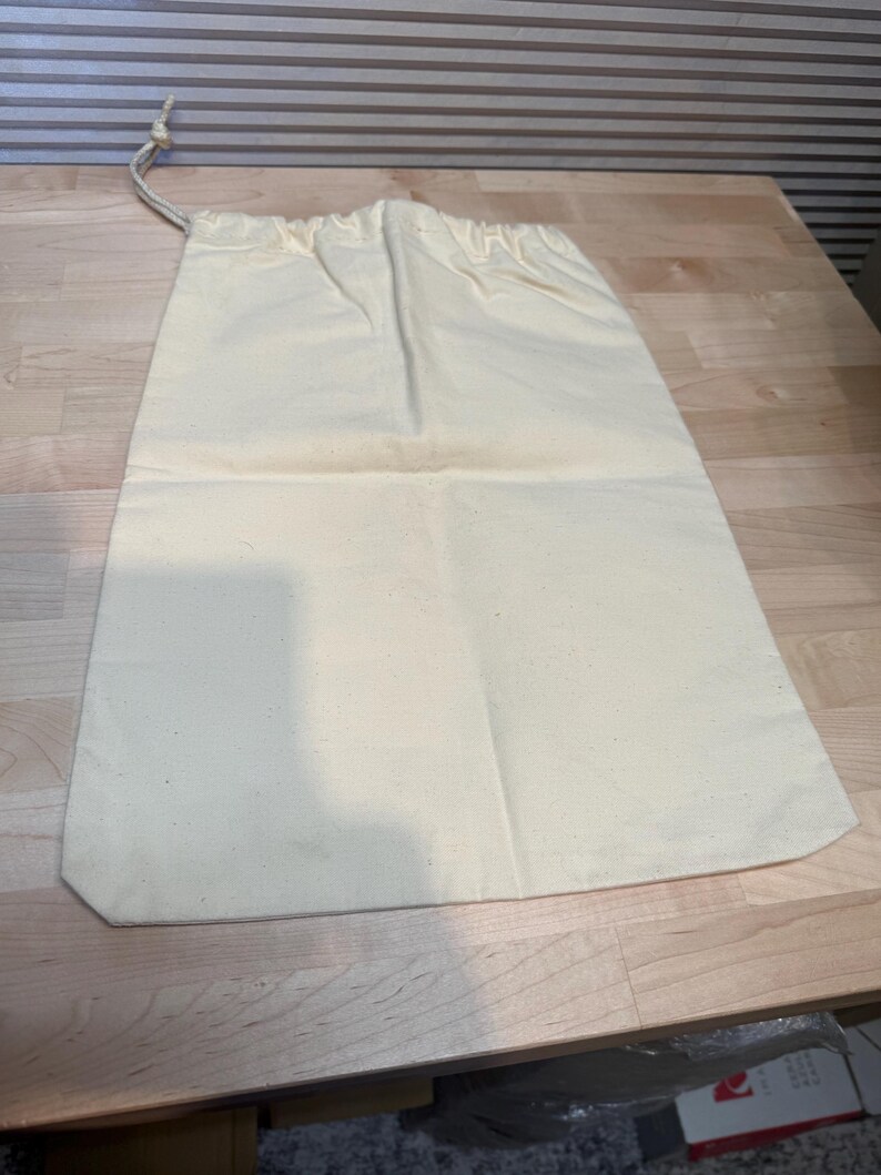 May include: A cream-colored drawstring bag made of canvas fabric. The bag is rectangular with a rounded bottom and a drawstring closure at the top. The bag is laying on a wooden surface.