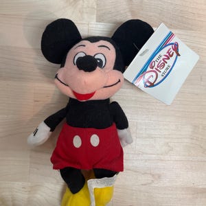 May include: A Mickey Mouse soft toy with black ears, a black body, red shorts with white buttons, yellow shoes, and white gloves. The toy has a tag that says "The Disney Store".